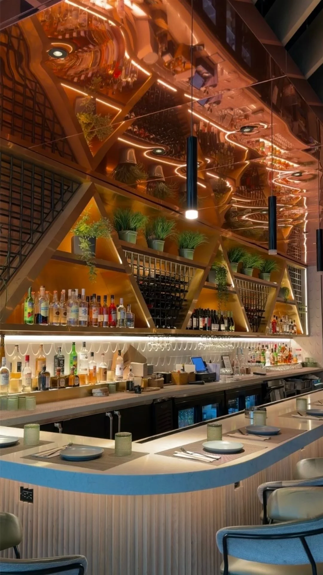 Interior of a modern bar with a mirrored ceiling, bottles of alcohol on shelves, green plants in triangle-shaped shelves, and table settings with plates and glasses.