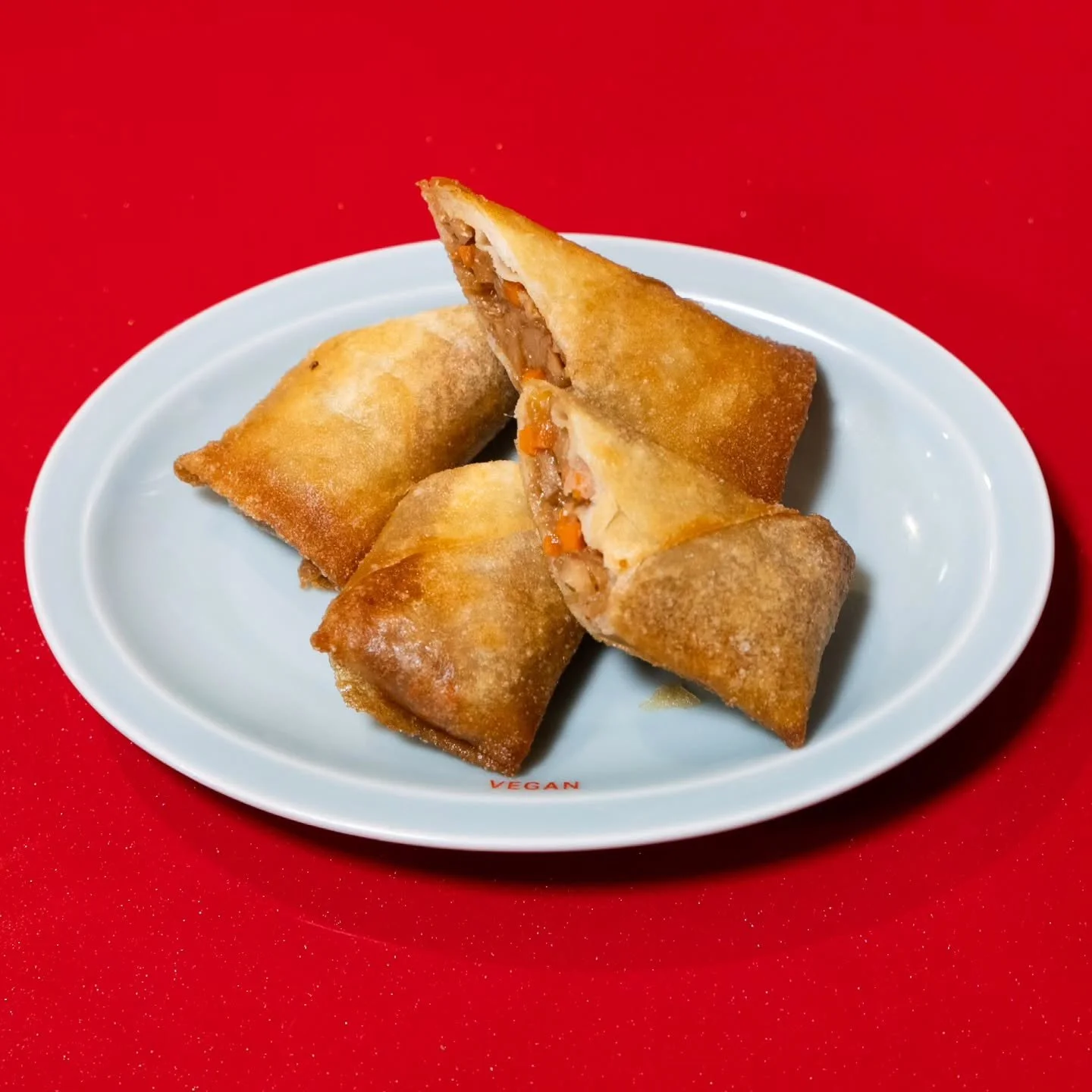 Four vegan spring rolls with a crispy golden exterior, one cut in half showing a vegetable filling, served on a white plate with red background