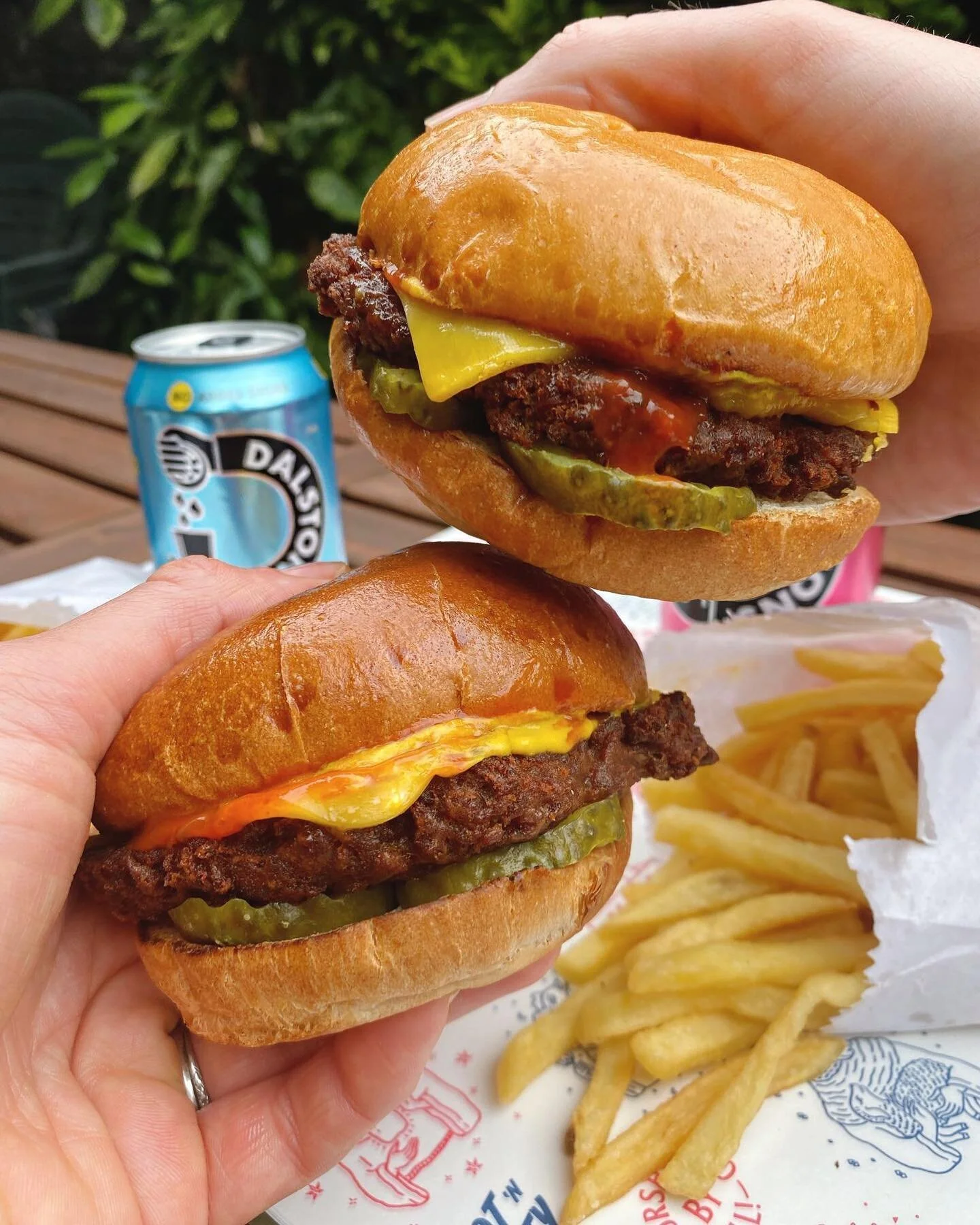 Two people holding cheeseburgers with pickles, cheese, and beef patties, along with a side of French fries on a paper tray, outdoors. In the background, a can of soda.