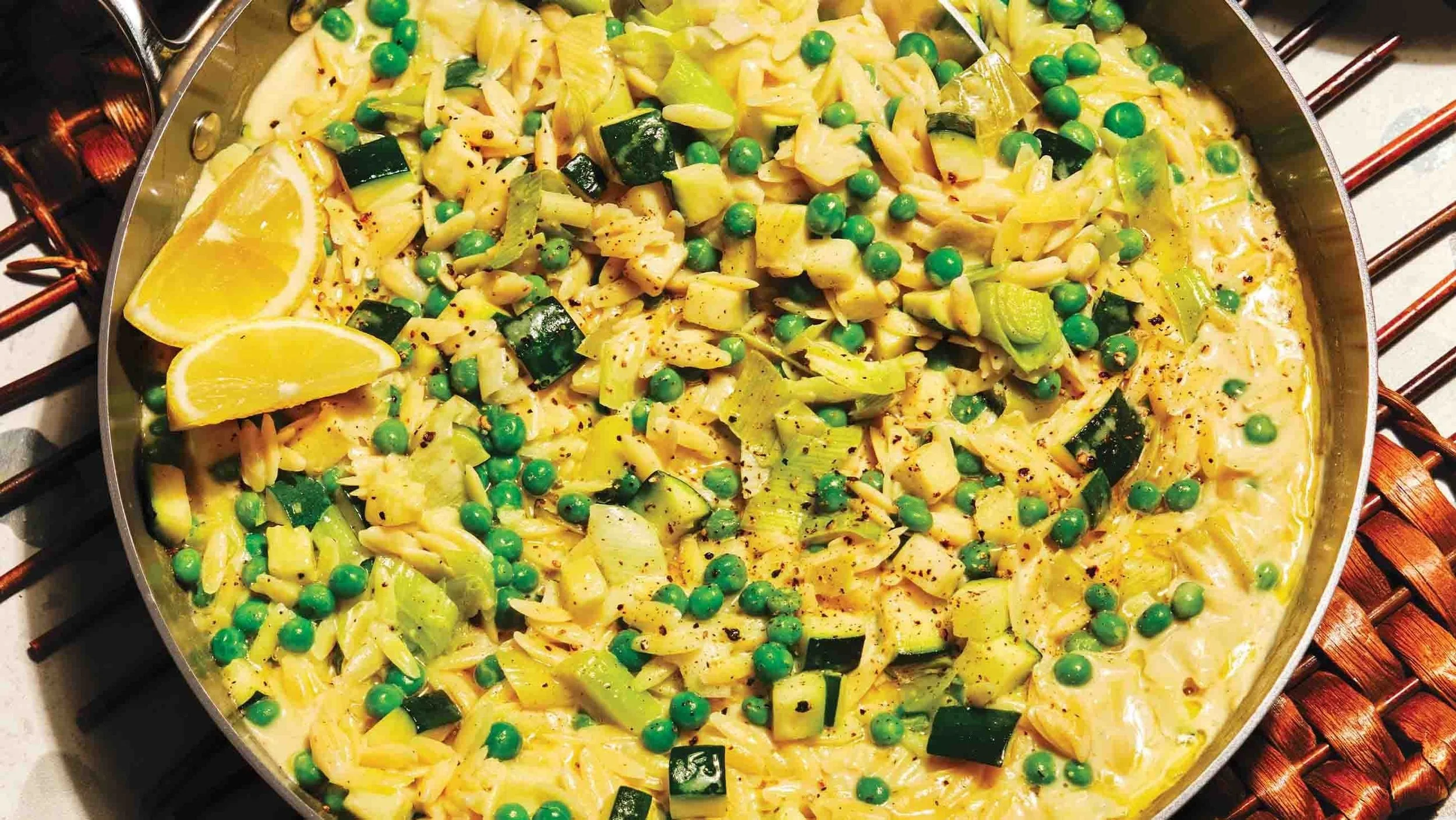 A skillet of cooked pasta with green peas, zucchini, and lemon wedges, seasoned with black pepper.