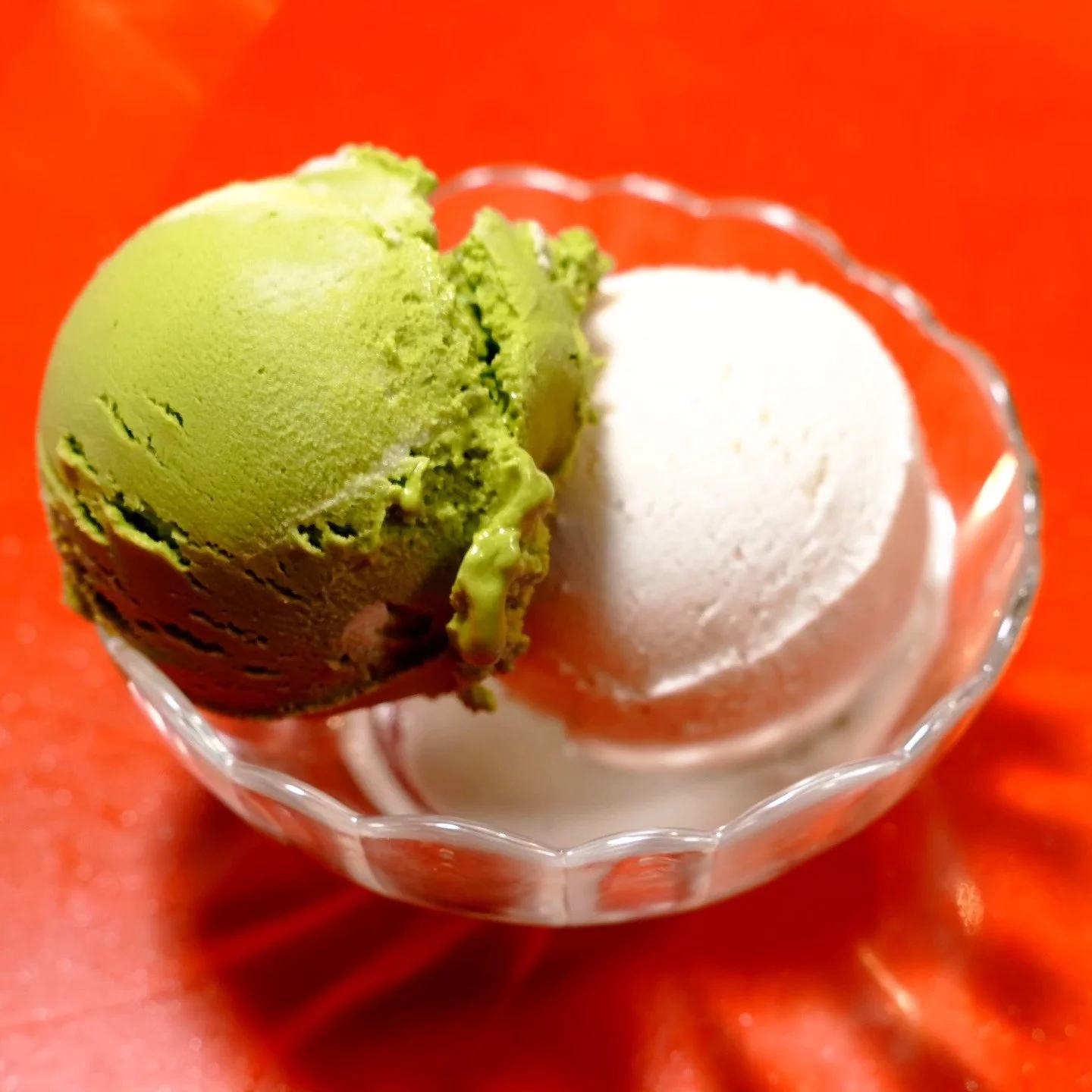 Two scoops of green and white ice cream in a small glass dish, on a red surface.