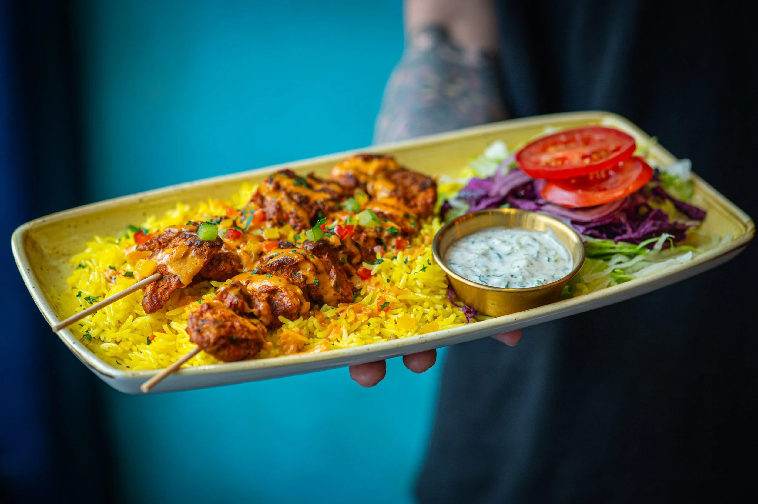 A person holding a yellow rectangular plate with Indian food, including yellow rice, grilled skewered meat, a small cup of creamy white sauce with herbs, and a side salad with sliced tomatoes, purple cabbage, and lettuce.