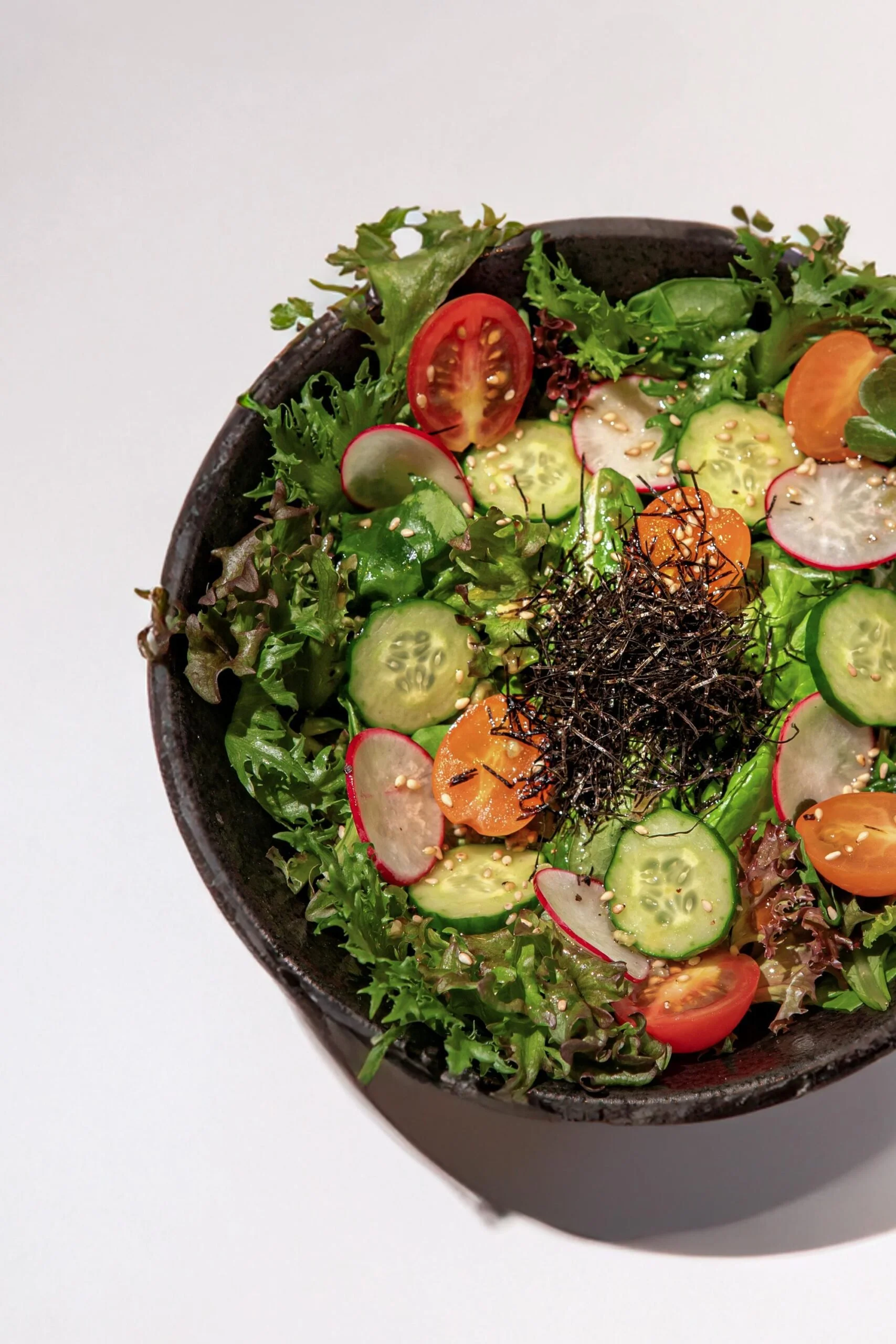 Close-up of a bowl of fresh salad with cherry tomatoes, cucumber slices, radish, mixed greens, and sprinkled sesame seeds.