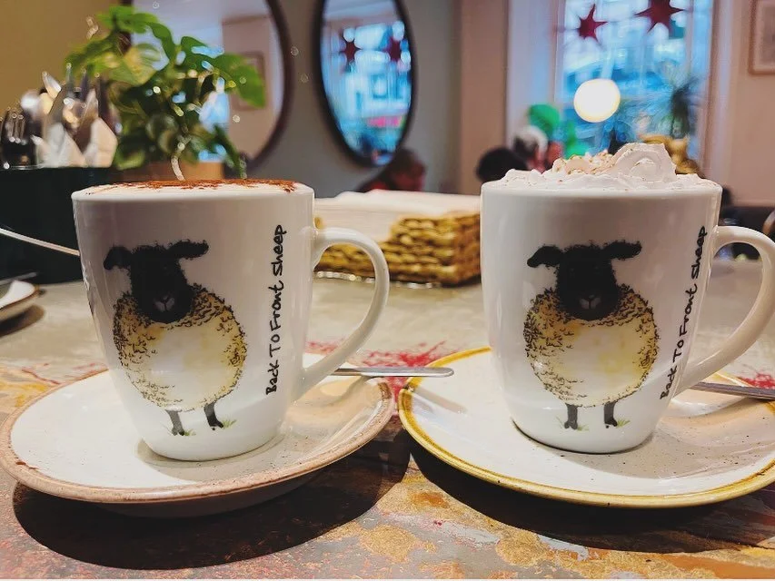Two coffee mugs with sheep illustrations and phrases, one topped with whipped cream, on a table in a cozy cafe.