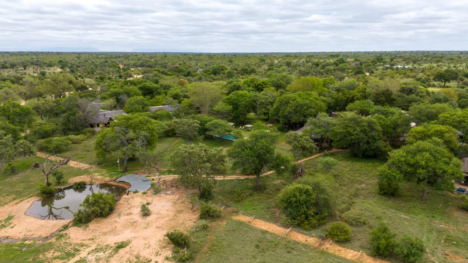 Aerial view of a lush, green landscape with scattered trees, small buildings, a pond, and dirt paths under a cloudy sky.