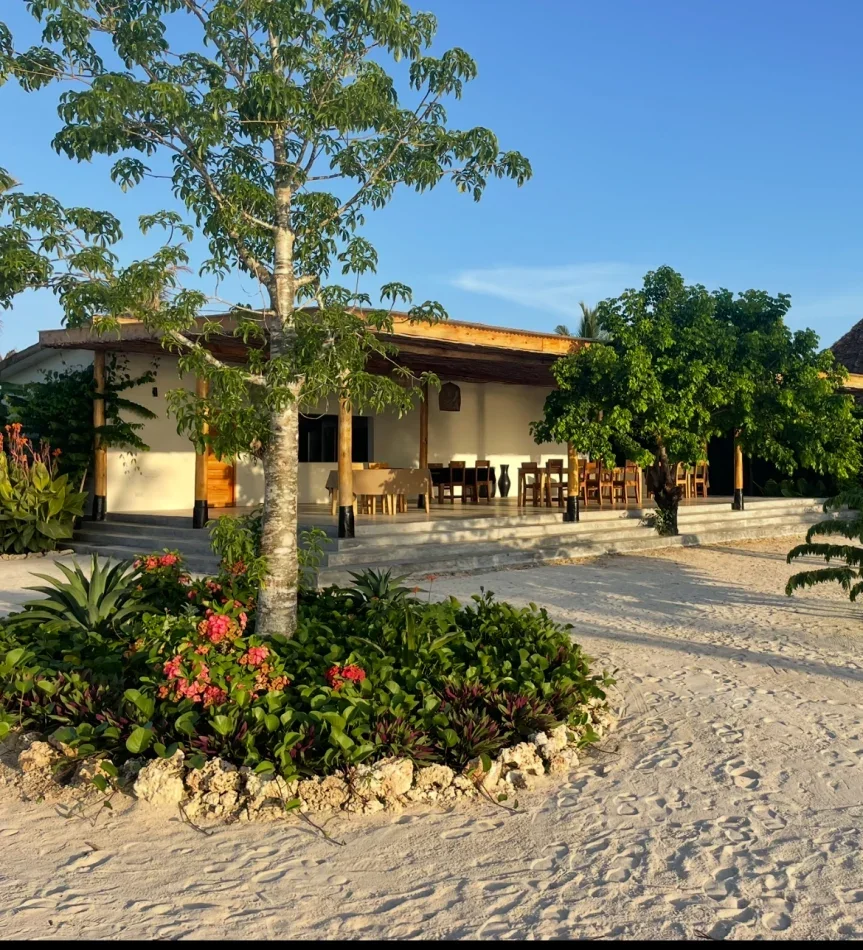 A beachside building with a porch, surrounded by green trees and flowering plants, on sandy ground with a clear blue sky.