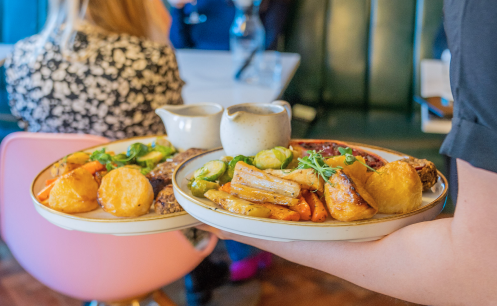 Person carrying two plates of food, including roasted vegetables and fried potatoes, in a restaurant setting.