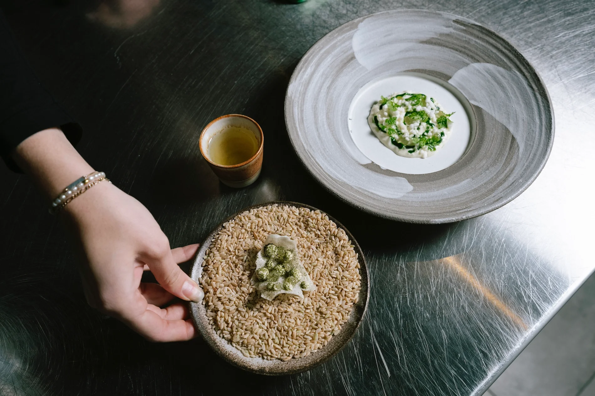 A person is holding a bowl of sunflower seeds topped with a dollop of white sauce or cream and sprinkled with green powder. On the table, there is a grey plate with a greenish dish garnished with herbs, a white dish with a creamy substance garnished 