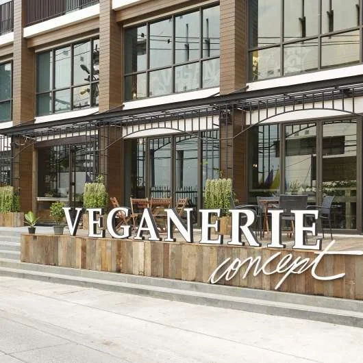 Exterior view of a vegan restaurant called Veganerí Concept with large glass windows, outdoor seating, and potted plants in front.