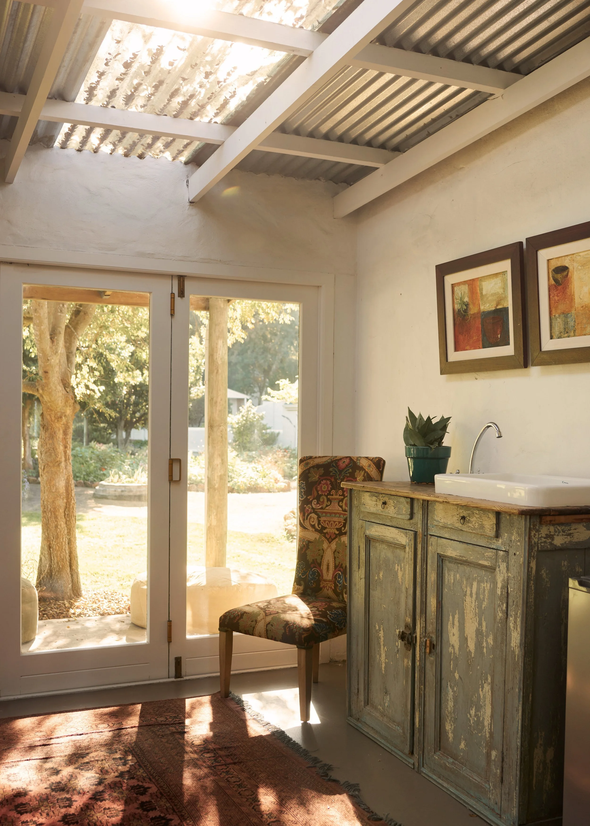 Sunlit room with a glass-paneled door leading to a garden, a vintage painted wooden cabinet with a sink, a patterned upholstered chair, framed artwork on the wall, and a potted plant on the cabinet.