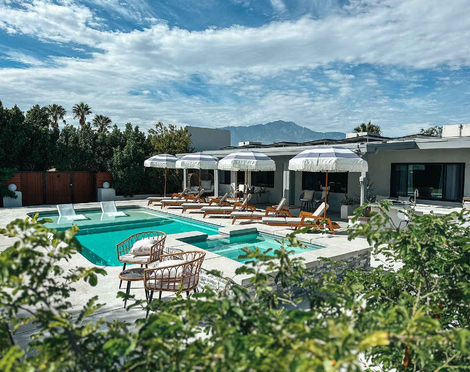 A backyard with a swimming pool surrounded by lounge chairs and umbrellas, with a modern house in the background and mountains in the distance under a partly cloudy sky.