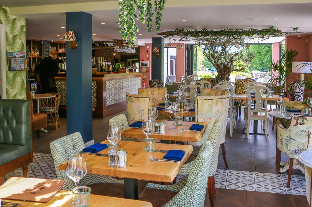 Elegant restaurant with wooden tables set with wine glasses, blue napkins, and cutlery, surrounded by patterned upholstered chairs, with a bar area and large window showing greenery outside.