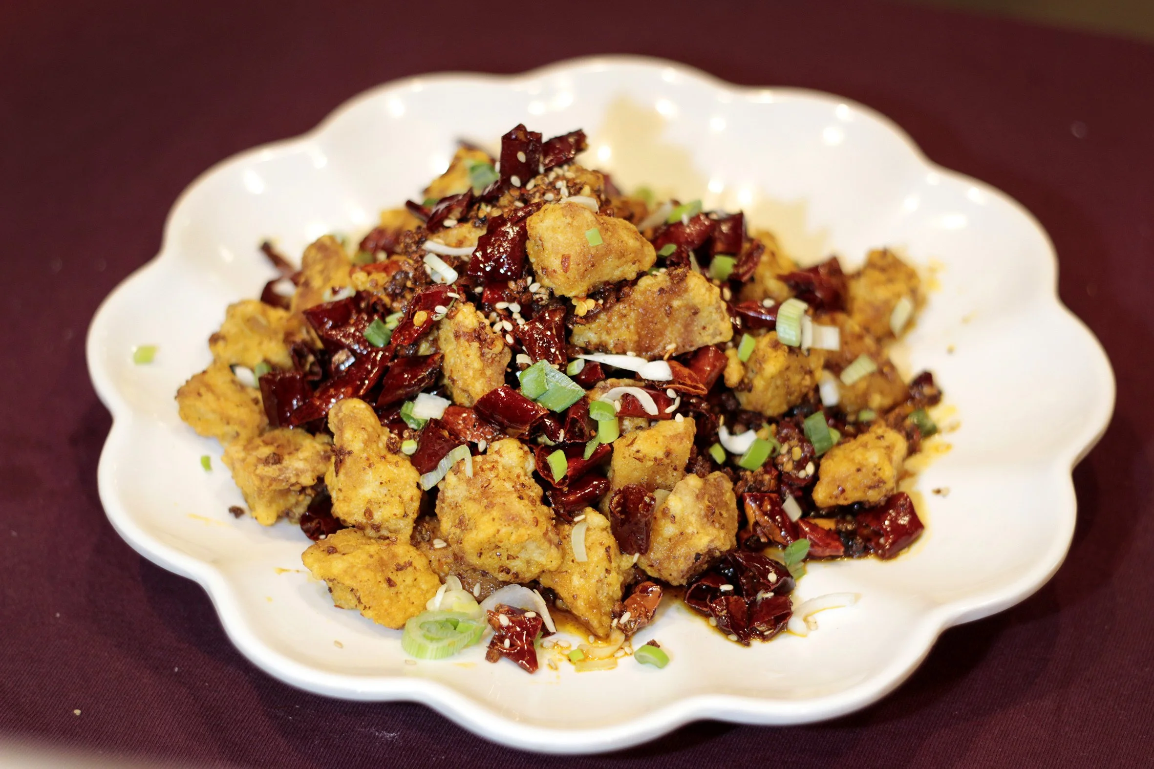 A white scalloped bowl filled with a spicy fried tofu dish topped with dried red chili peppers, chopped green onions, and sesame seeds, placed on a dark red surface.