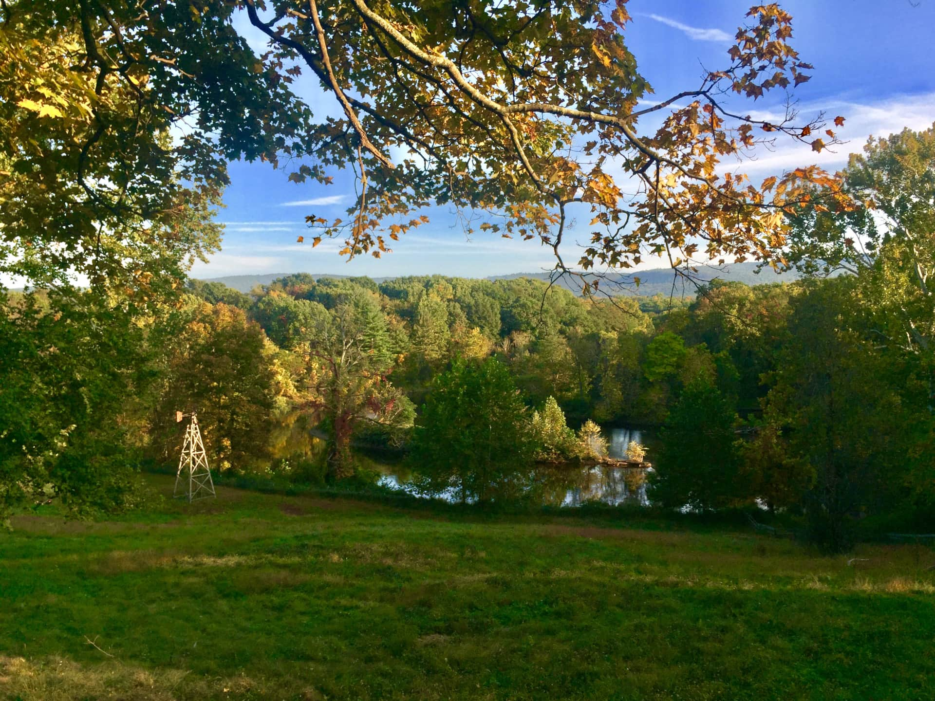 A scenic landscape of a river surrounded by trees with green, orange, and yellow leaves, under a clear blue sky with some clouds, during autumn.