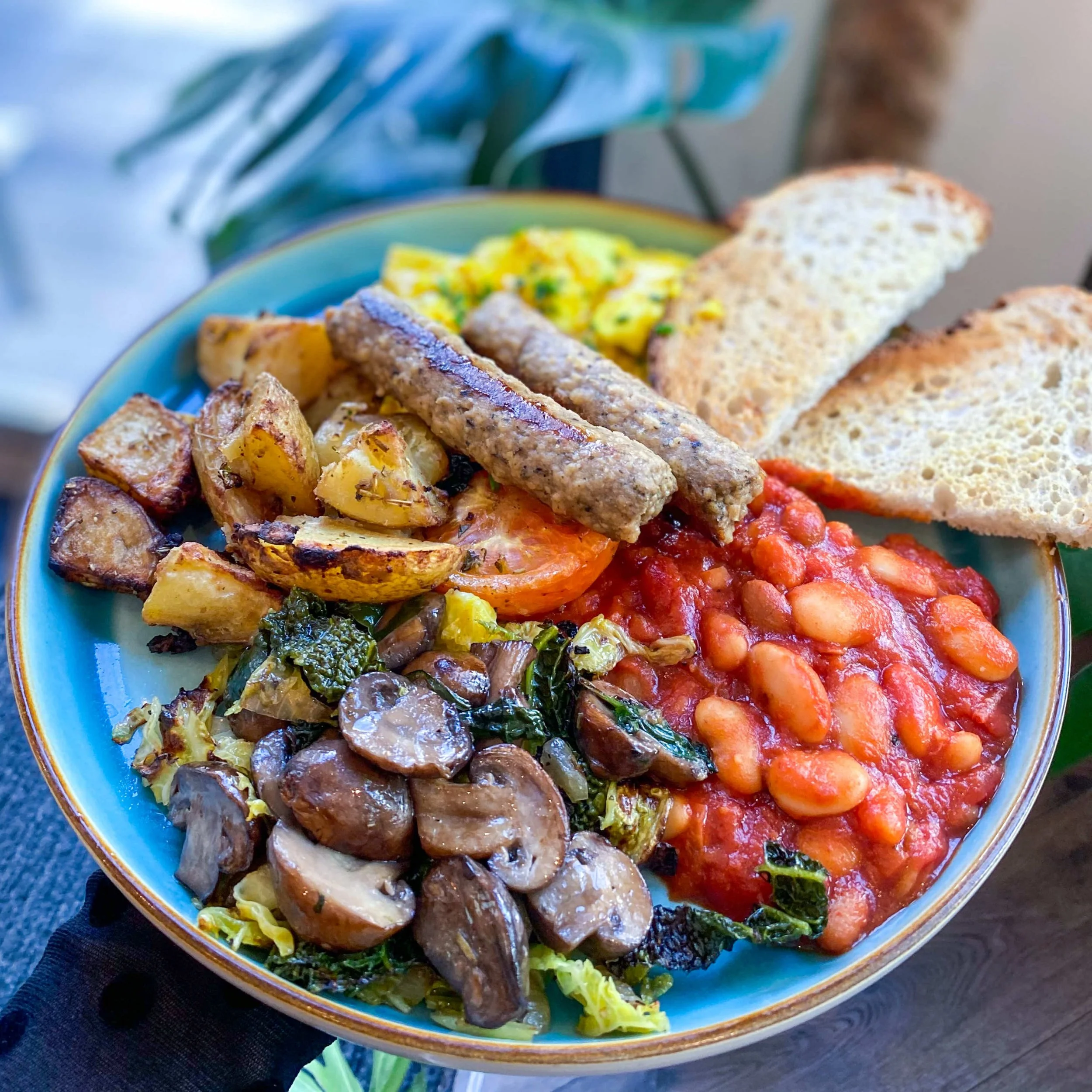 A colorful breakfast plate with baked beans, sautéed mushrooms and greens, roasted potatoes, scrambled eggs with herbs, two sausages, and slices of toasted bread.