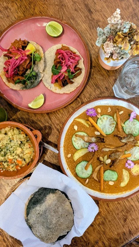 A meal featuring two tacos with pink pickled onions, a lime wedge, a bowl of rice, a dish with curry and vegetables garnished with flowers, and a fried breaded item on paper, all on a wooden table.