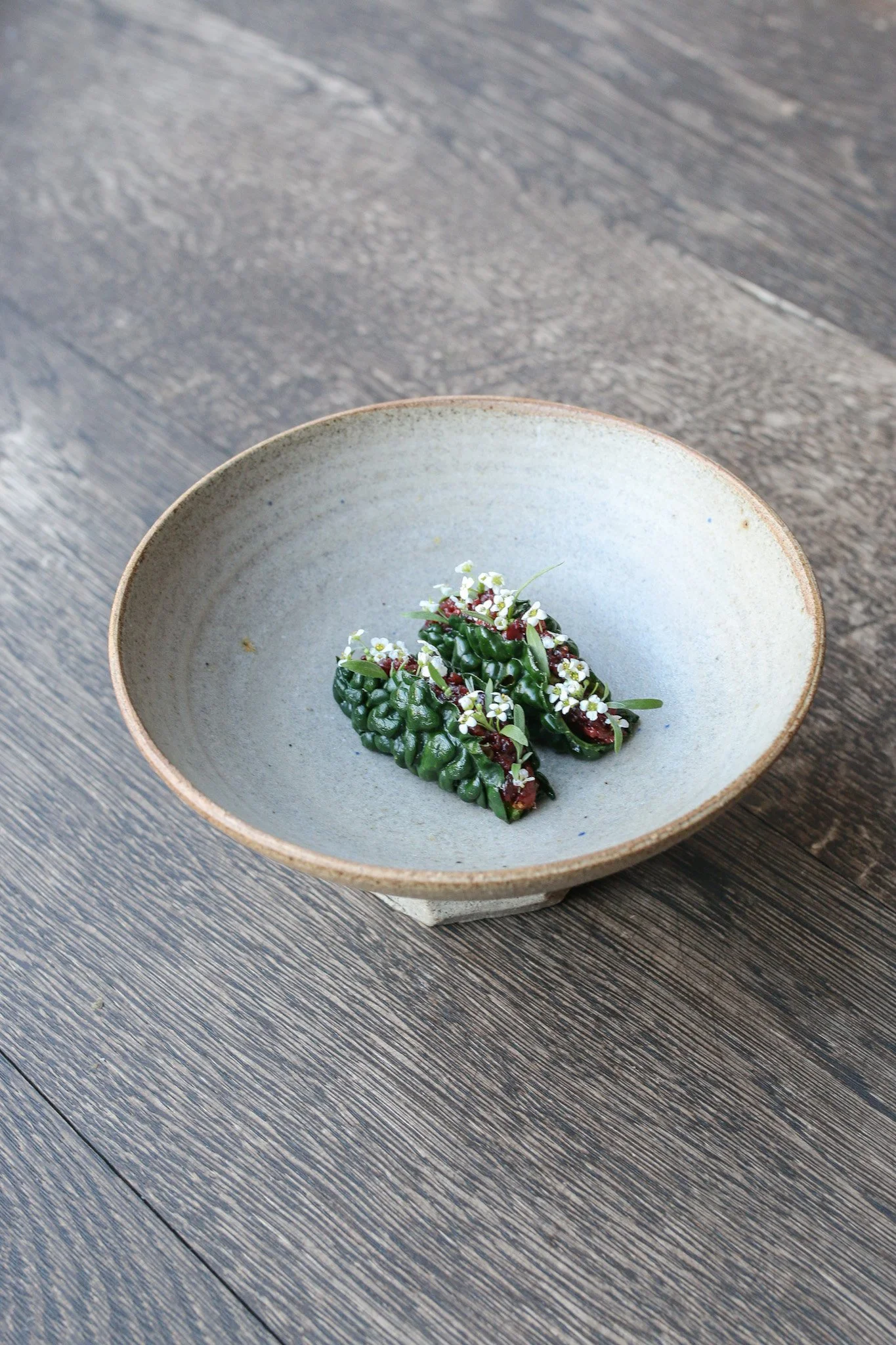 A ceramic bowl with a textured, matte finish contains a small arrangement of green leaves, tiny white flowers, and red berries, placed on a dark wooden surface.