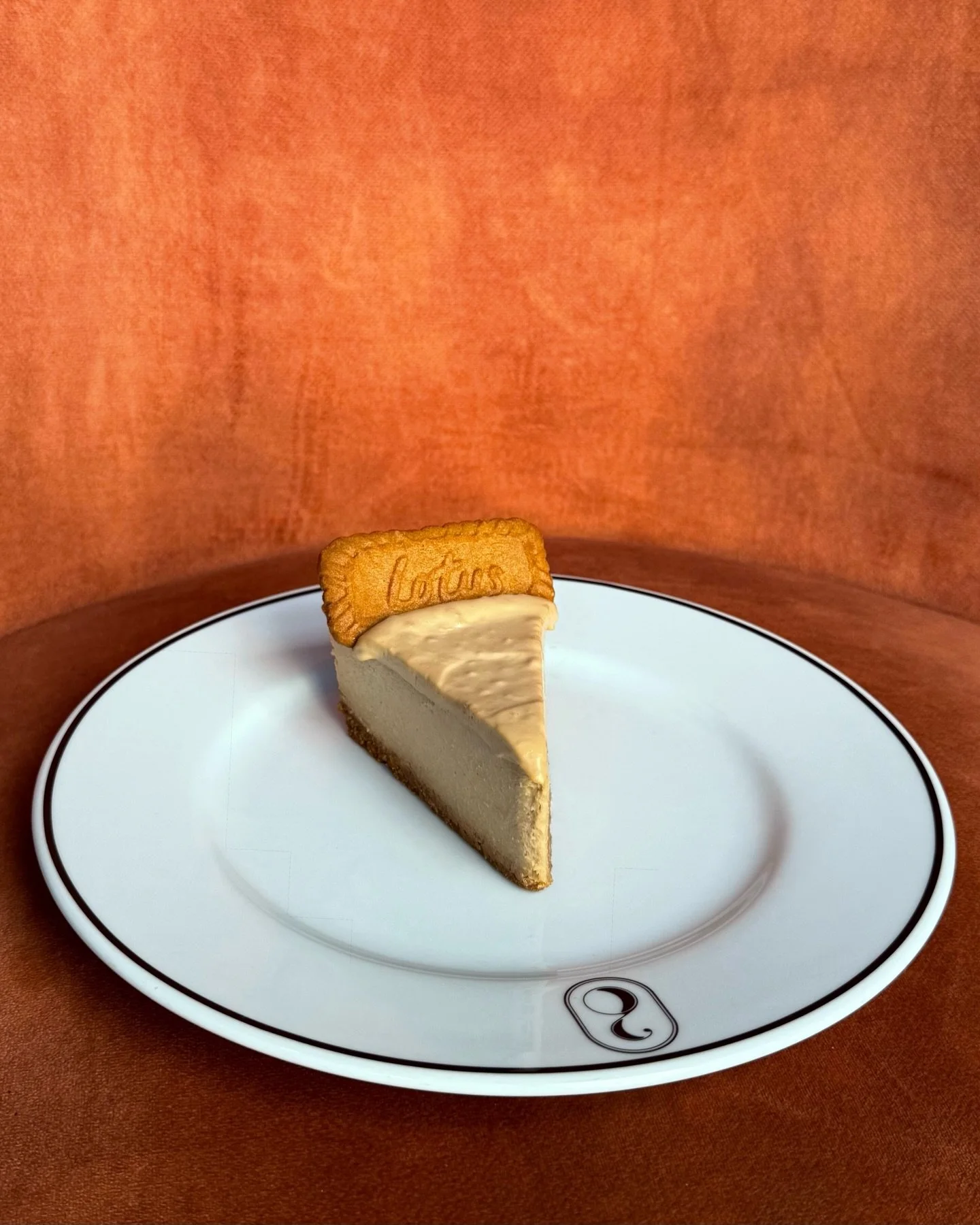 A slice of cheesecake with a biscuit decorated with the word 'Lottus' on top, served on a white plate with a black trim.