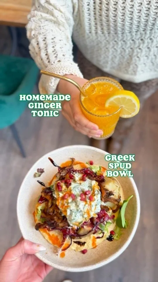 A person holding a glass of homemade ginger tonic with a lemon wedge, and another person holding a Greek spud bowl topped with vegetables, sauce, and pomegranate seeds.
