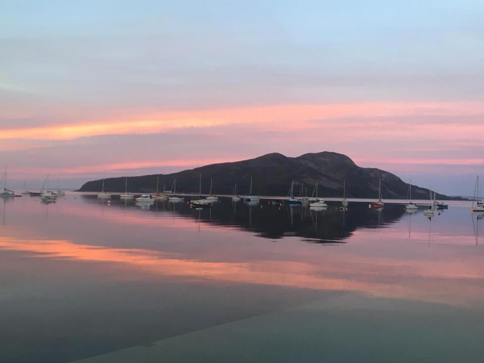 A tranquil scene of sailboats anchored on calm water with a mountainous island in the background during sunset, reflecting pink and blue hues in the water.
