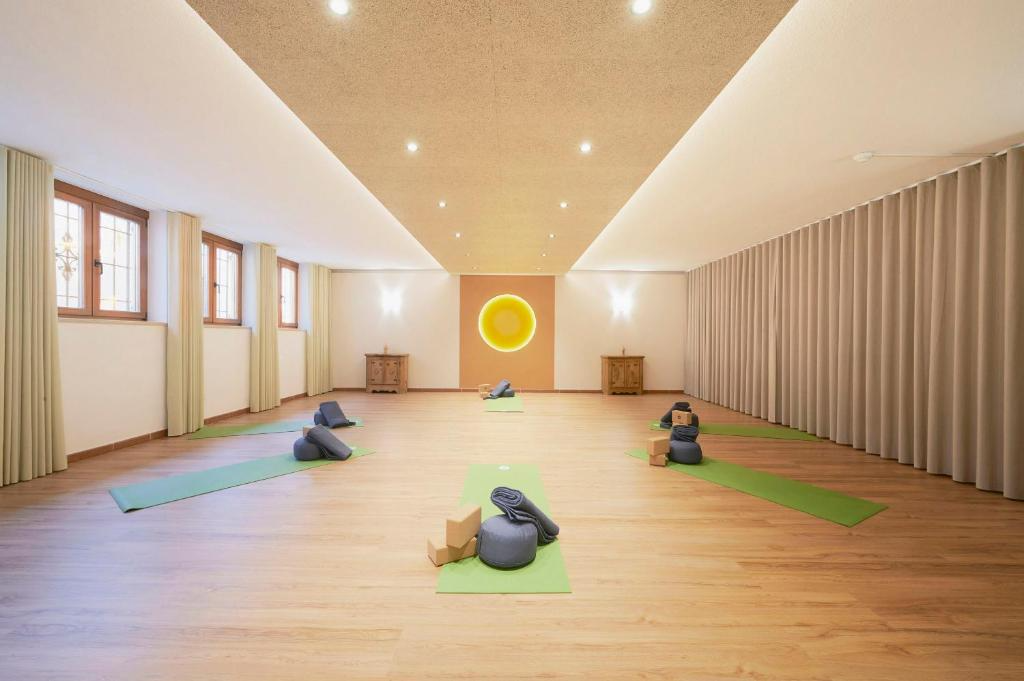A spacious yoga studio with wooden floors, four green mats with yoga props, beige curtains, and soft lighting. There is a glowing yellow sun decoration on the back wall.