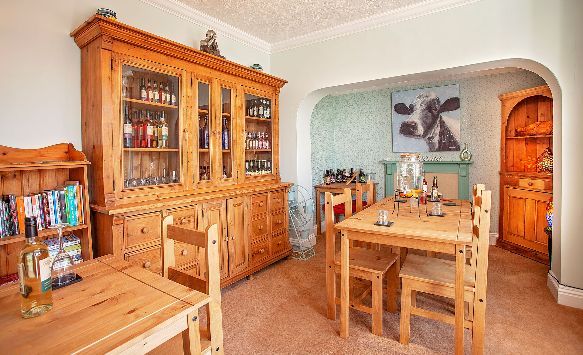 A cozy dining room with wooden furniture, including a table, chairs, and cabinets, decorated with books, bottles, glasses, and a large painting of a cow on the wall.