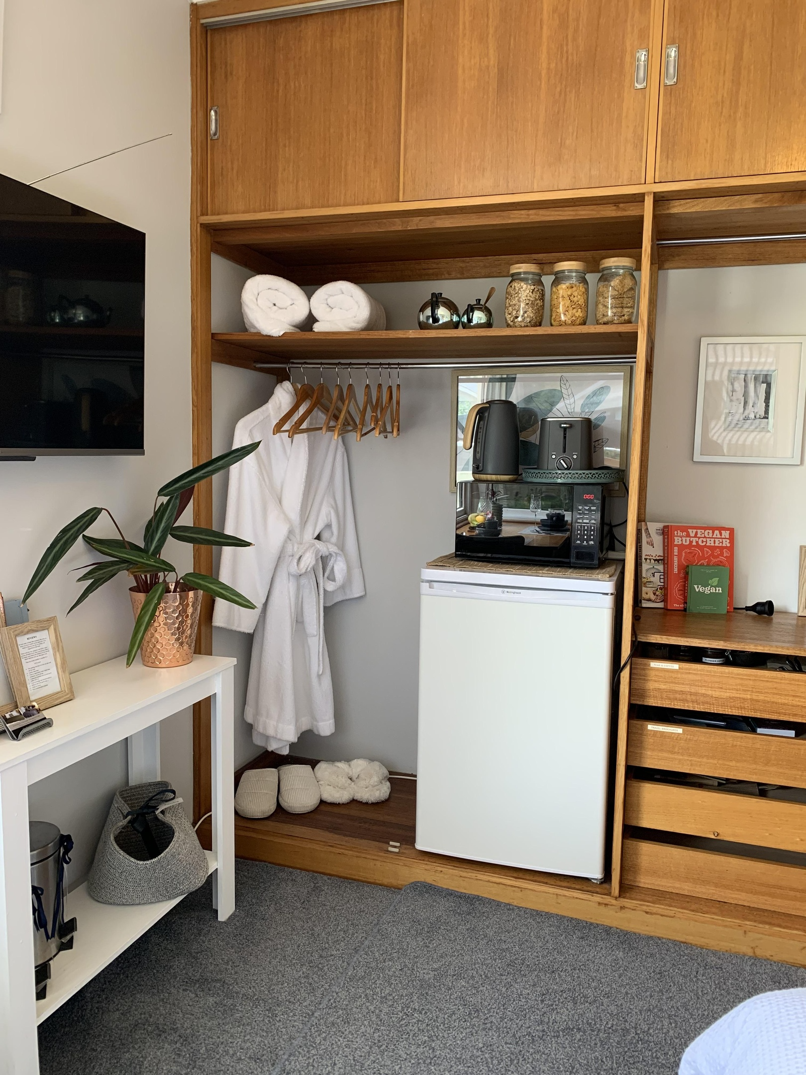 A cozy corner of a room with wooden shelving, a small white fridge, a microwave, a kettle, jars of cereal, a framed watercolor art, books, and slippers on the floor. There is a white bathrobe hanging and a grey carpeted floor with plants nearby.