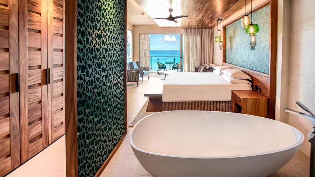 Interior of a beachside hotel room with a freestanding bathtub, a large bed, seaside balcony with chairs, wood-paneled ceiling, and modern lighting.
