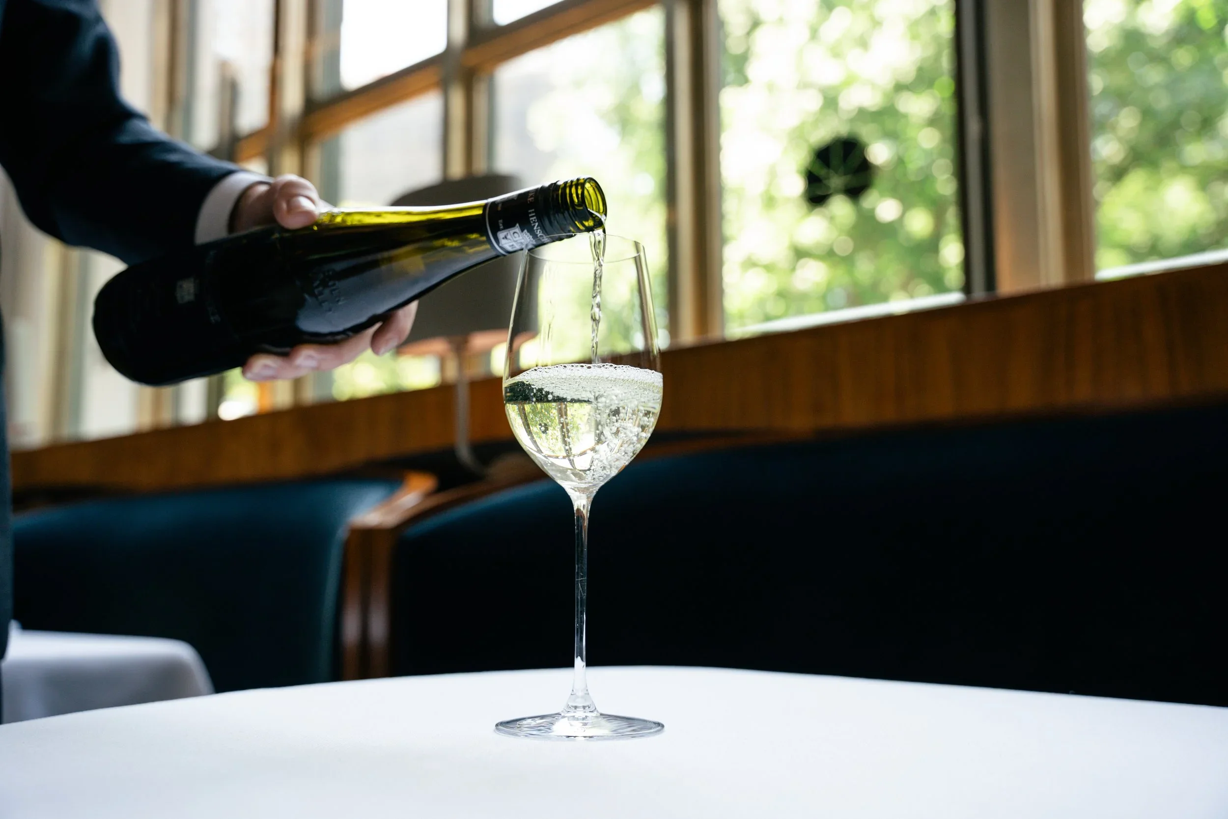 A hand pouring sparkling wine into a glass on a white table in a well-lit room with large windows and green foliage outside.