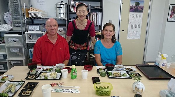Three people in a dining setting with Japanese food on the table. Two are seated, and one woman stands behind them, smiling.
