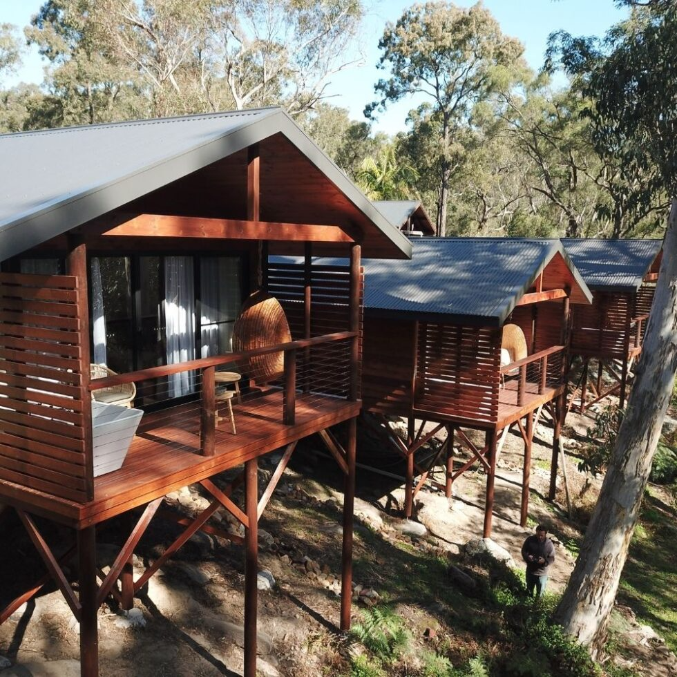 Wooden stilt cabins in a wooded area with trees in the background, featuring outdoor balconies and metal roofs.