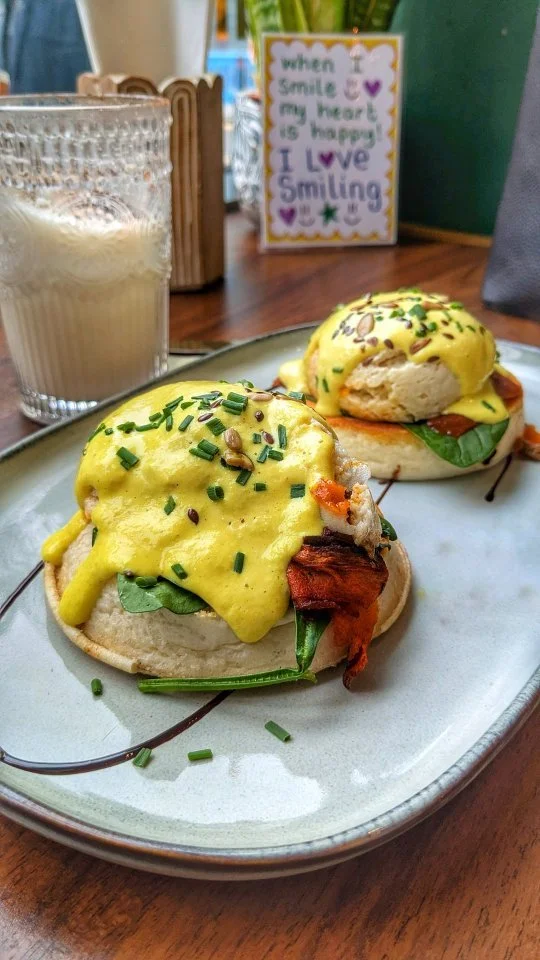 Two breakfast sandwiches topped with yellow eggs, chives, and seeds, served on a ceramic plate. A glass of iced coffee and a colorful decorative sign with a positive message are in the background.
