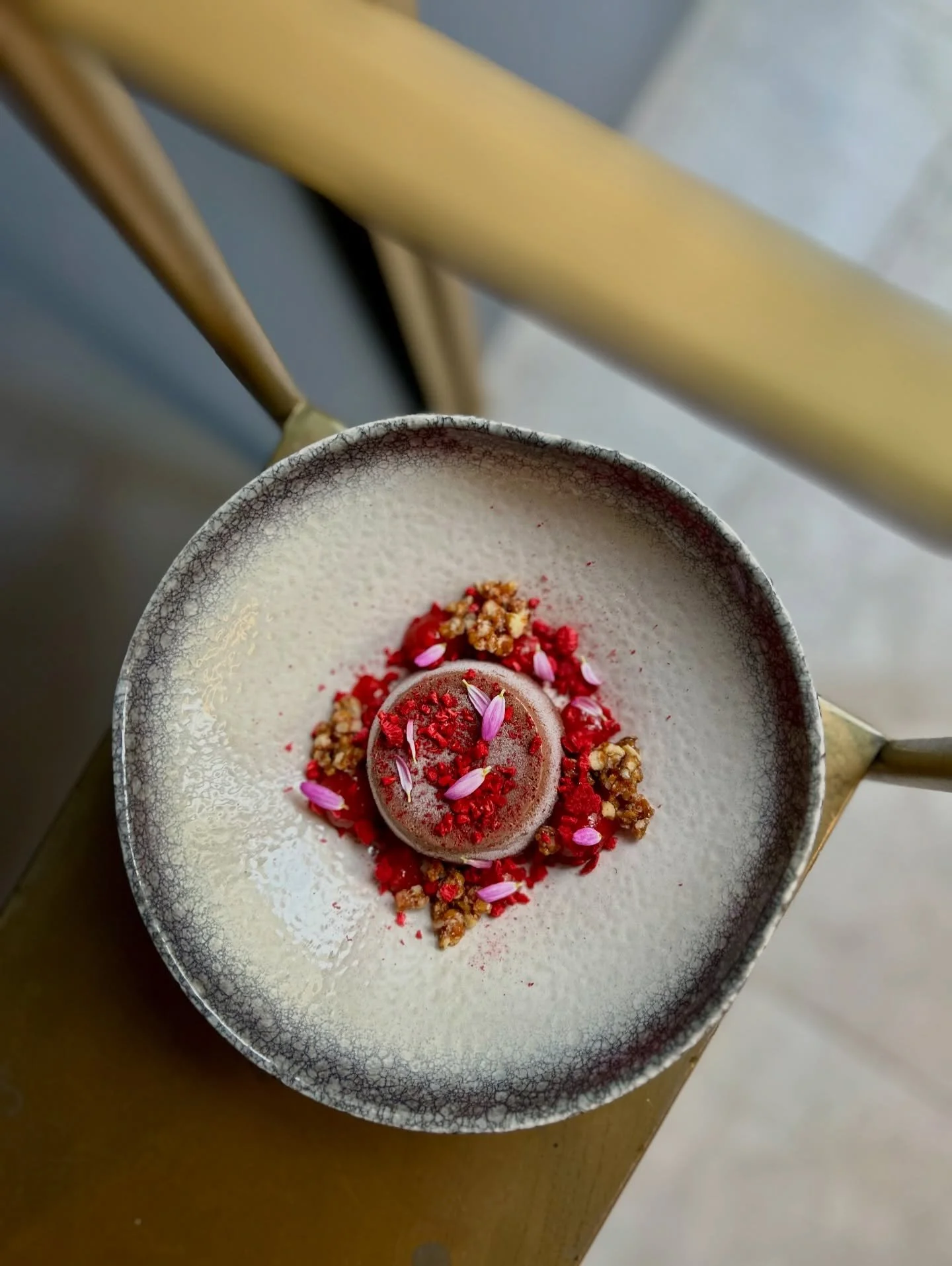 A dessert on a speckled white plate, garnished with red dried berries, edible pink flower petals, and small crunchy nut pieces.