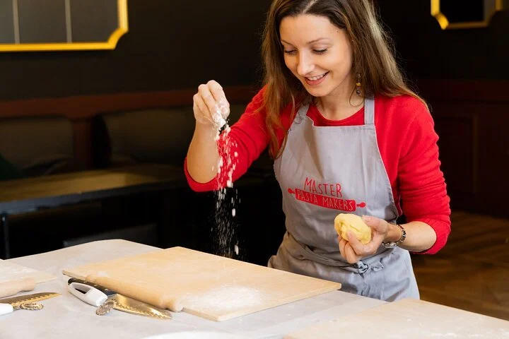 Young woman baking, sprinkling flour on a wooden surface in a kitchen, wearing a gray apron with red text and a red long-sleeve shirt.