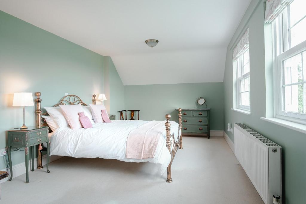 A bedroom with a mint green accent wall, a brass bed with white bedding and pink pillows, two matching bedside lamps, a dresser with a round mirror, and three large windows with white blinds, casting natural light.