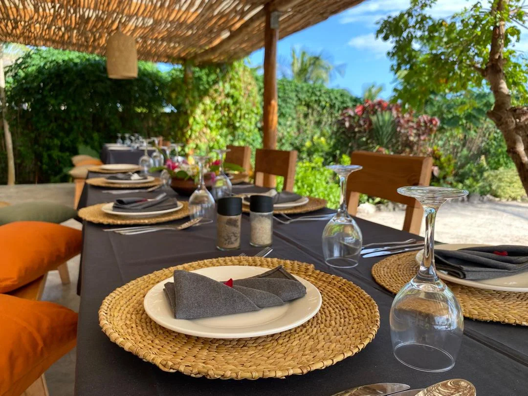 Outdoor dining table set with black tablecloth, white plates, gray napkins, wine glasses, and salt and pepper shakers. The table is under a thatched roof with lush green plants and trees in the background, suggesting a garden or patio setting.