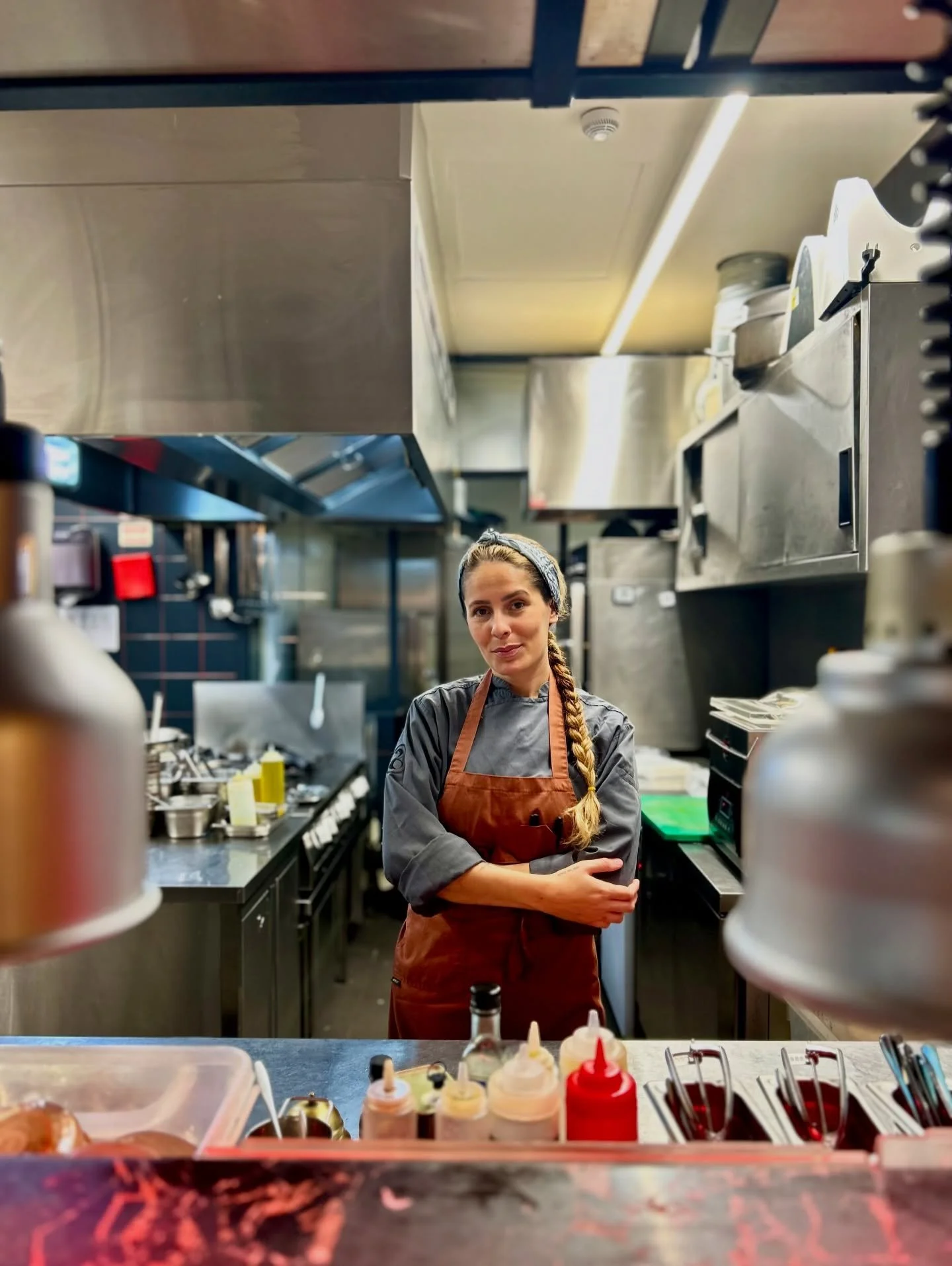 A female chef standing in a commercial kitchen with stainless steel appliances and counters, wearing a gray chef's coat and a brown apron, with a braid and headband, looking at the camera.