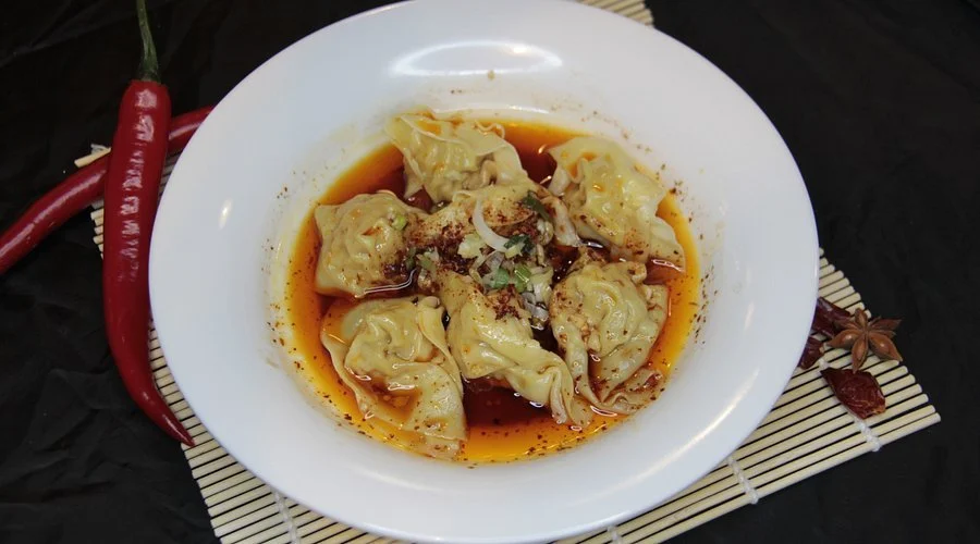 A white bowl of shumai dumplings in broth garnished with green onions and spices, with red chili peppers and star anise on a bamboo mat.