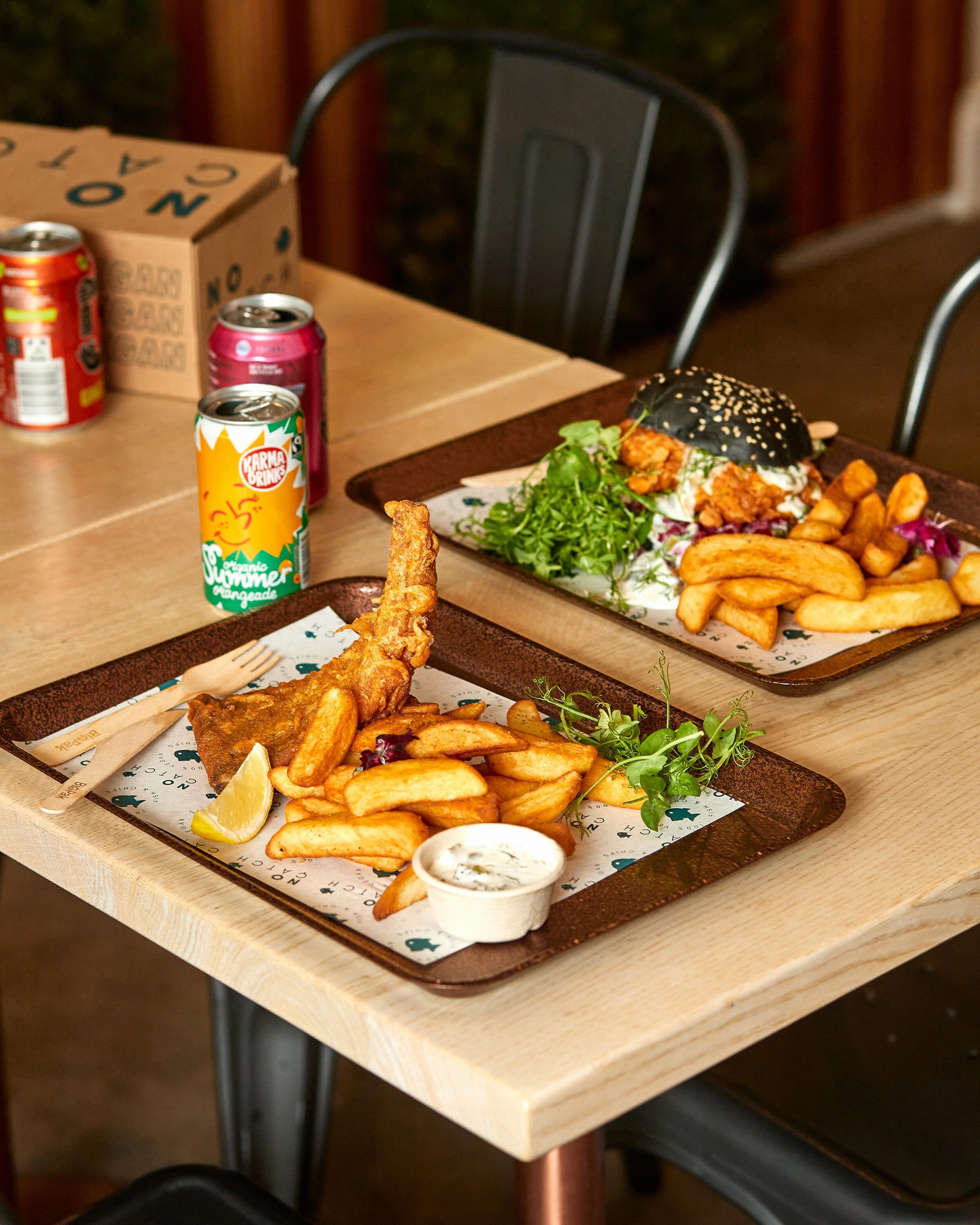 A meal on a dark tray with French fries, fried fish, dipping sauce, and lemon wedges at a restaurant table.