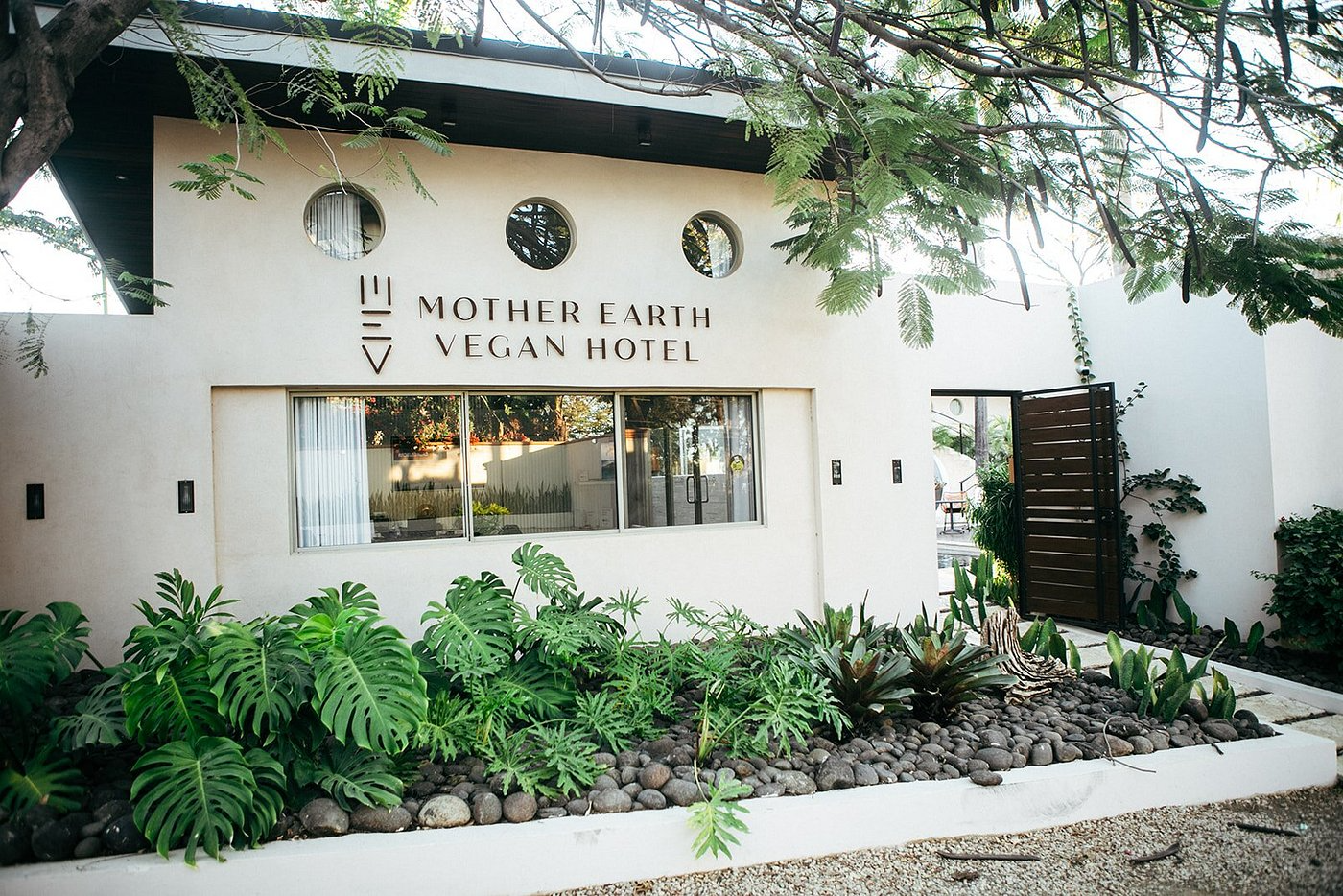 Exterior of Mother Earth Vegan Hotel with greenery and plants in front and a white building with circular windows.