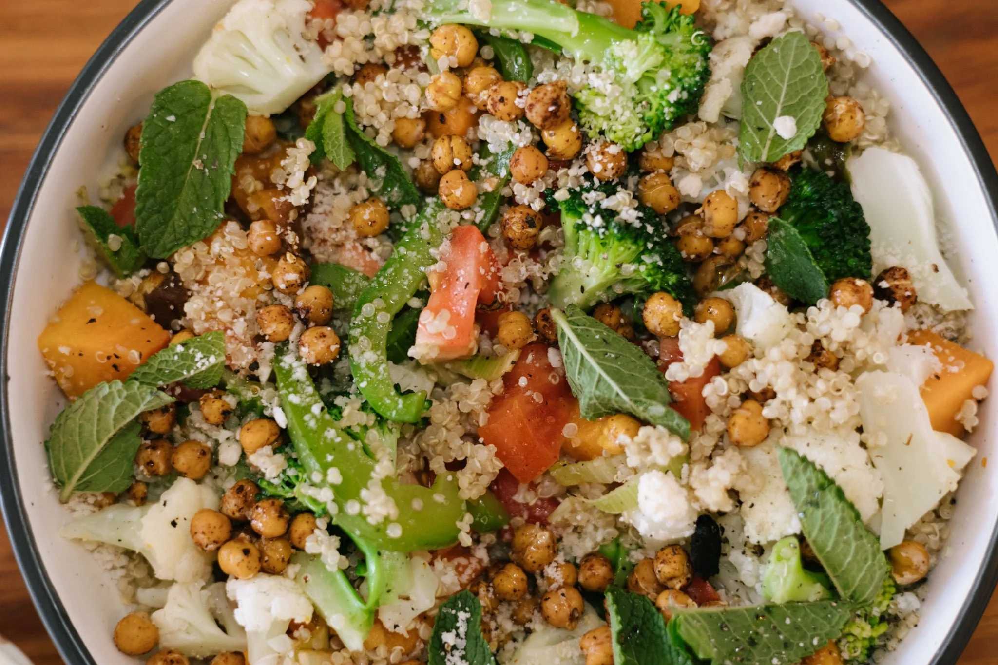 Close-up of a bowl containing a colorful salad with chickpeas, cauliflower, broccoli, carrots, fresh mint leaves, and some grated cheese.