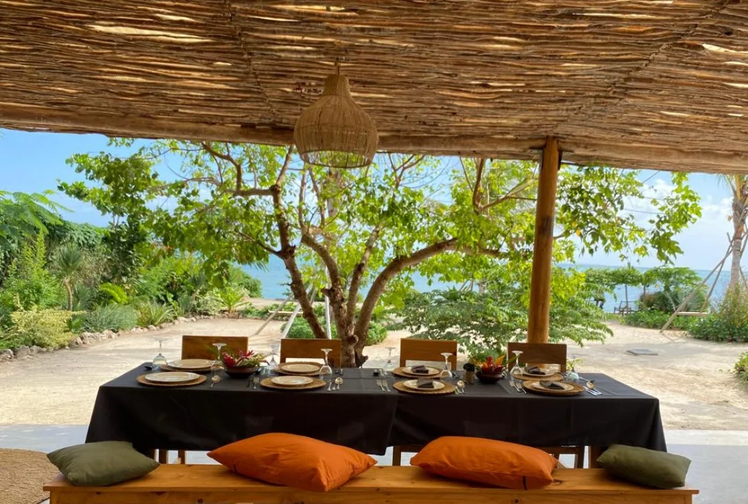 Outdoor dining area with a long table set with plates, glasses, and bowls, under a thatched roof with a view of trees and the ocean in the background.