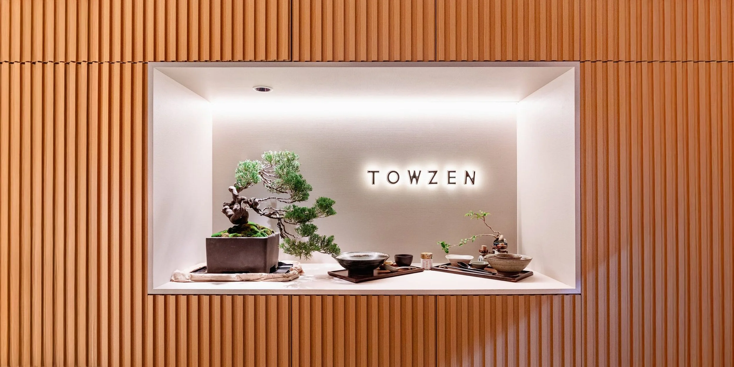 Display of a bonsai tree with various ceramics on a raised surface, with the word 'TOWZEN' illuminated on the wall behind, framed by vertical wooden slats.