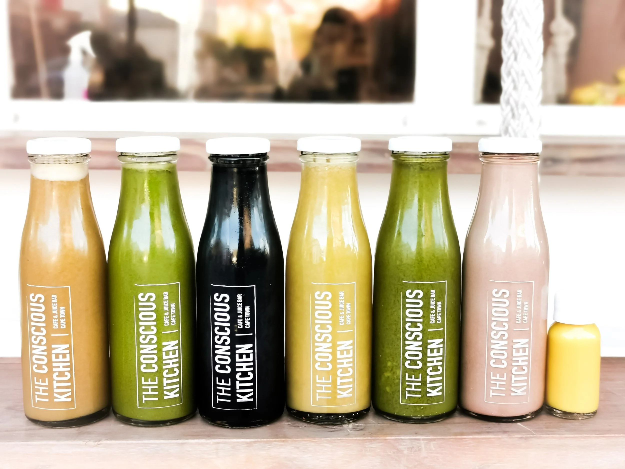Six bottled drinks lined up on a shelf, each with white caps and different colored liquids including beige, green, black, yellow, and pink, with a blurred background.