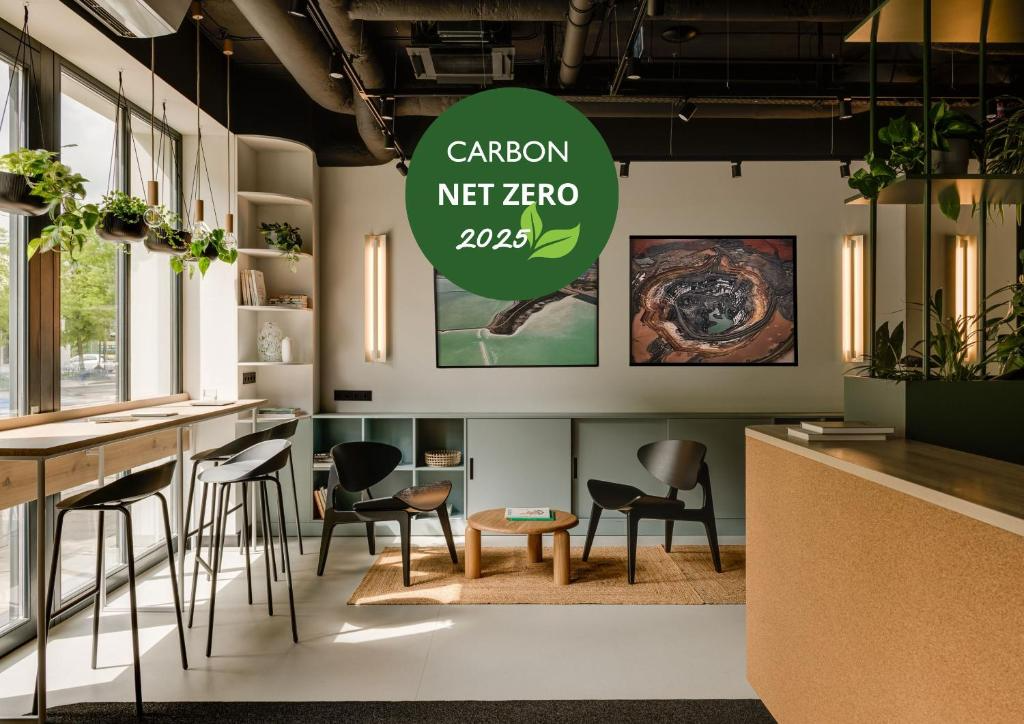 Modern office or lounge space with large windows, hanging plants, black chairs, a small round table, and framed artwork on the wall. Green sticker reads 'Carbon Neutral Zero 2025'.