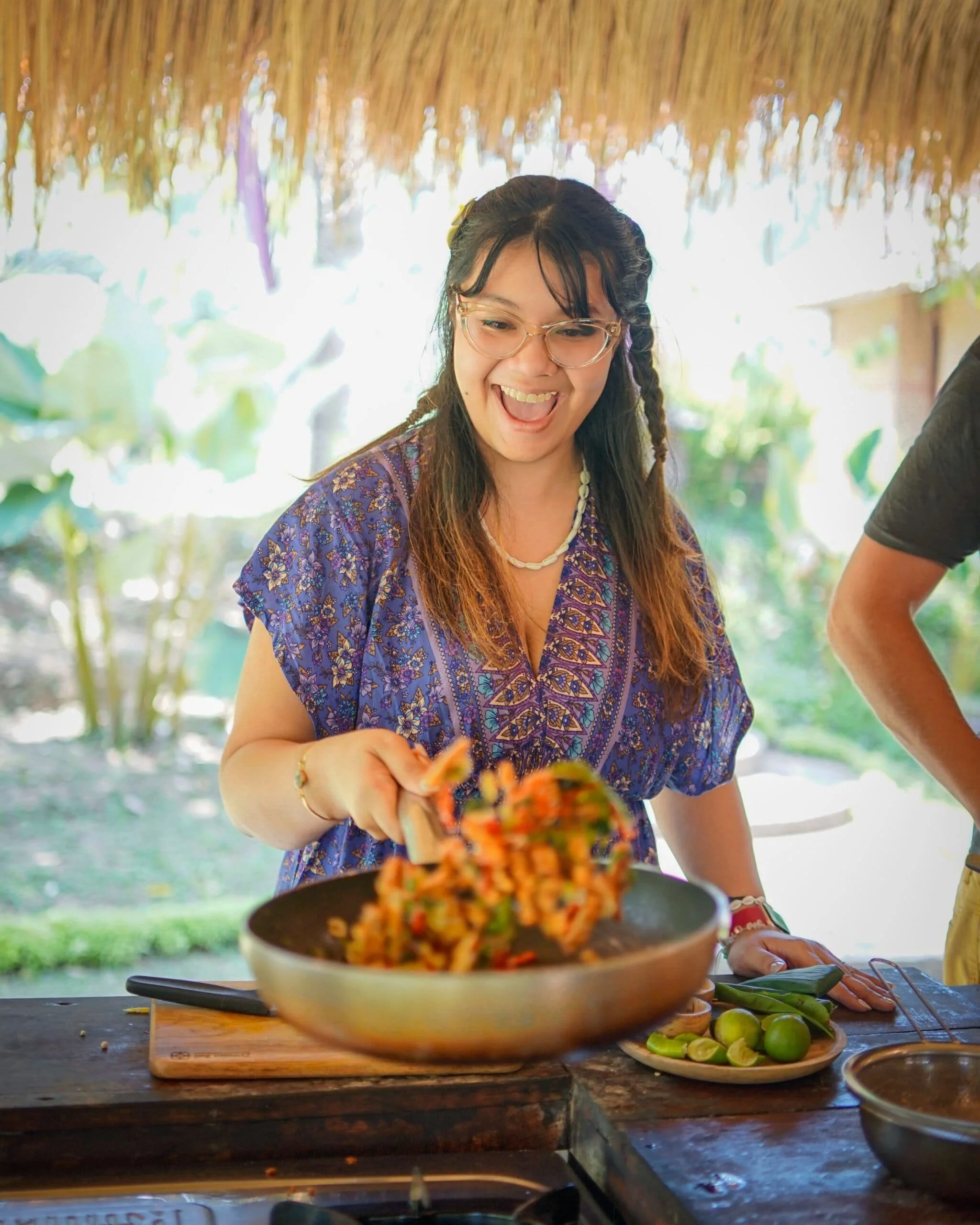A woman with glasses, dark hair, and a floral shirt is preparing food under a thatched roof. She is smiling and holding a spatula, with a plate of sliced limes and another dish nearby on the wooden table.