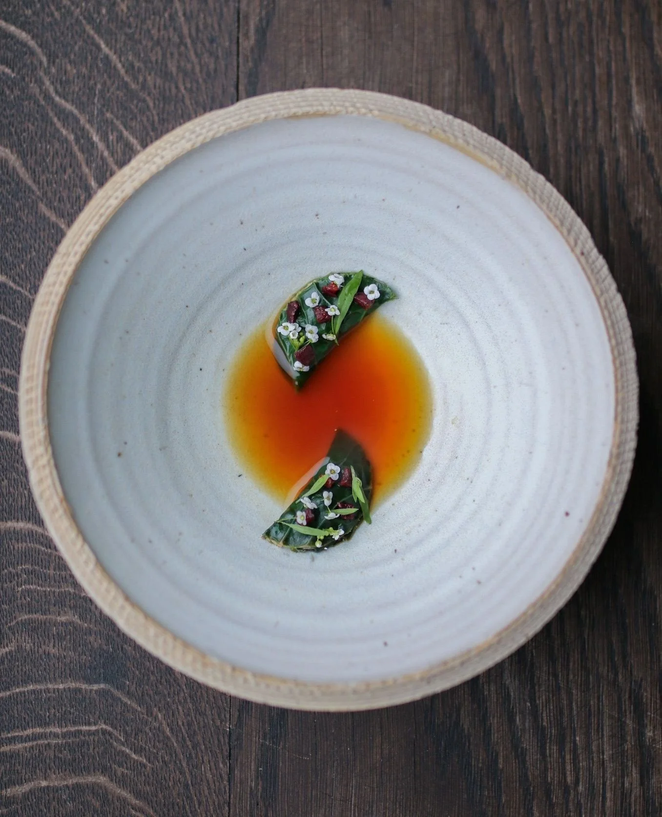 A white oval ceramic dish with a textured rim, containing a small, artfully arranged appetizer with green leaves, red toppings, tiny white flowers, and a reddish-binger sauce or broth at the bottom, placed on a wooden table.