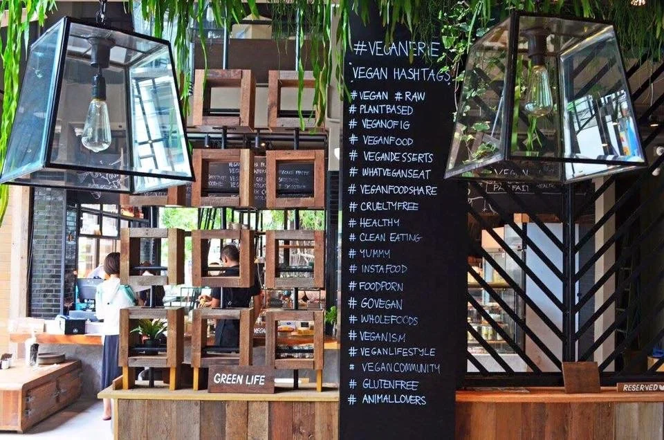 Interior of a vegan restaurant with a blackboard listing vegan hashtags, glass light fixtures, wooden shelves, and greenery.