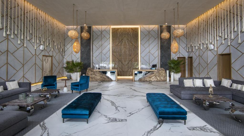 Modern hotel lobby with marble floors, gold accent walls, and unique hanging light fixtures. Contains black and blue velvet furniture, large potted plants, and a reception desk with natural stone accents.