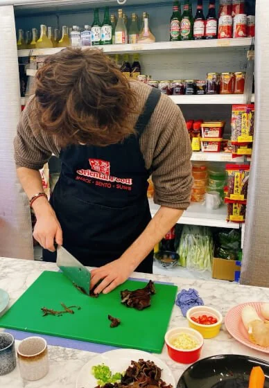Woman chopping herbs on a green cutting board in a grocery store kitchen with shelves of sauces and condiments in the background.